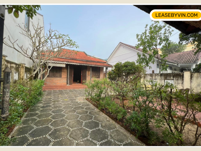 Hoi An A brick house with a red-tiled roof is at the end of a paved pathway bordered by green shrubs and trees. The entrance to the house includes a small porch. Next to the house, the large yard is enclosed by walls covered with vines. The sky is clear. Text: LEASEBYVIN.com.