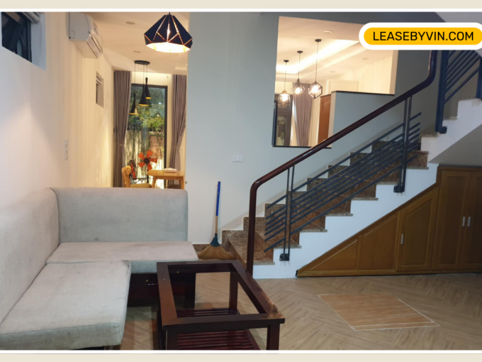 Hoi An A modern living area in a 3-bedroom house features a light gray sectional sofa and a wooden coffee table. To the right is a staircase with a metal railing, leading upstairs. In the background, a dining area is visible with a large window. A "LEASEBYVIN.com" logo is in the upper right corner.