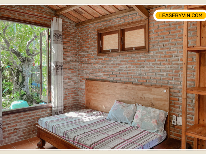 Hoi An Cozy bedroom with a brick wall, wooden ceiling, and large window offering a view of lush greenery. The bed features a plaid bedspread and decorative cushions. A wooden shelf on the right enhances the rustic charm, giving it an auto draft of natural warmth.